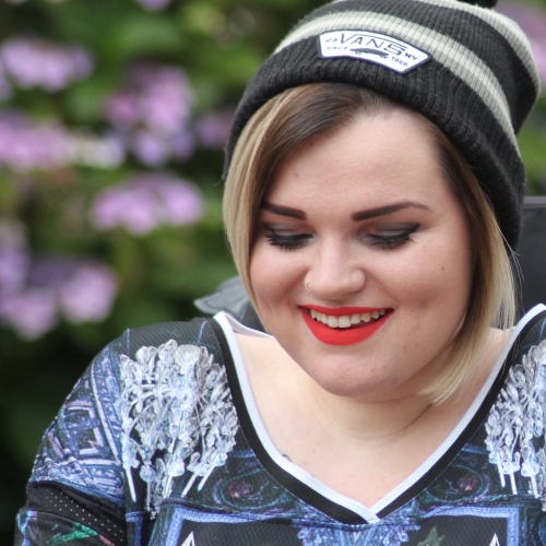 Kelly Perks-Bevington smiling downwards wearing a beanie hat and purple hydrangeas blurred in the background