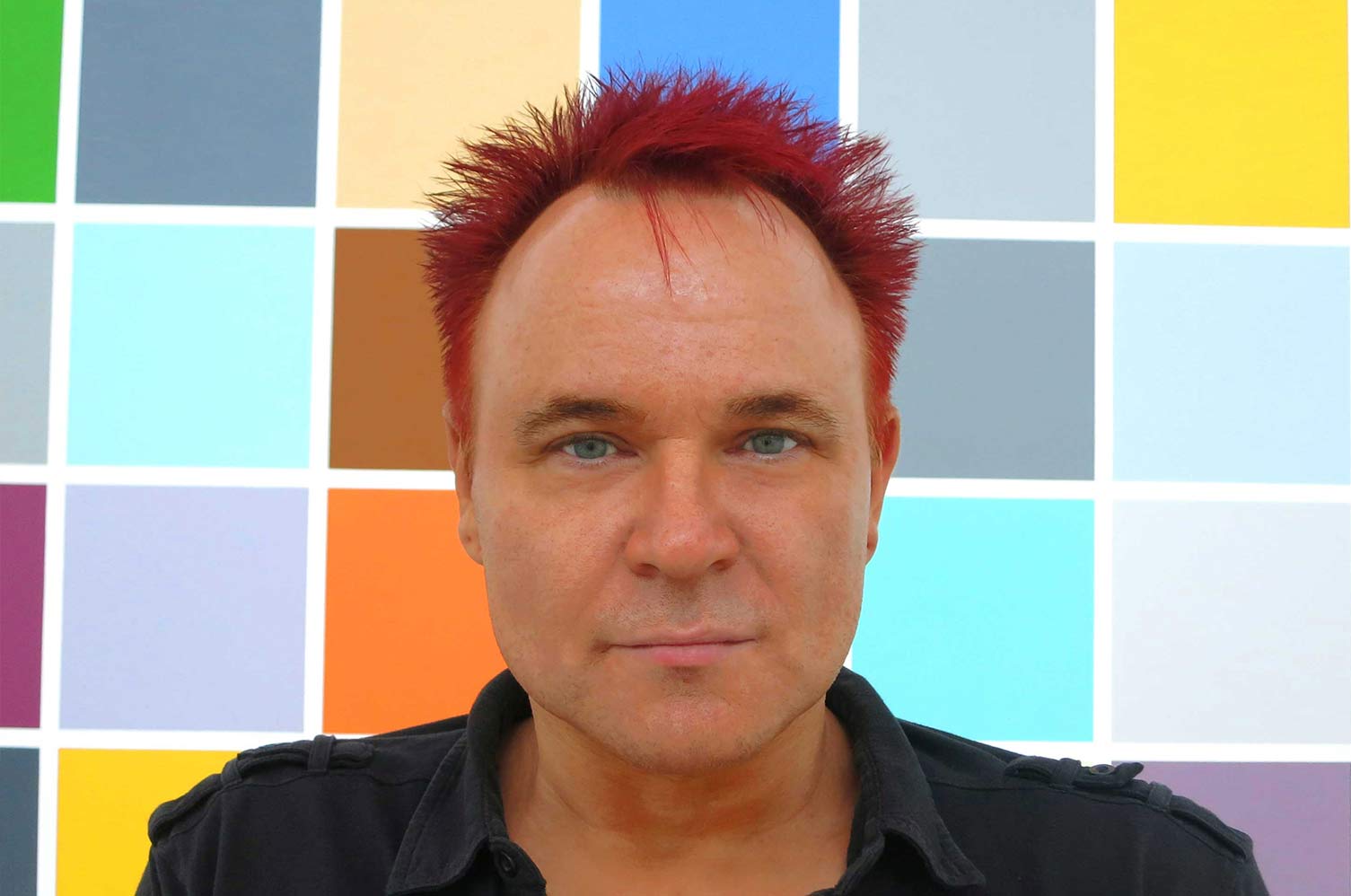 Mik looking directly into the camera, with red spikey hair and a coloured grid pattern behind him