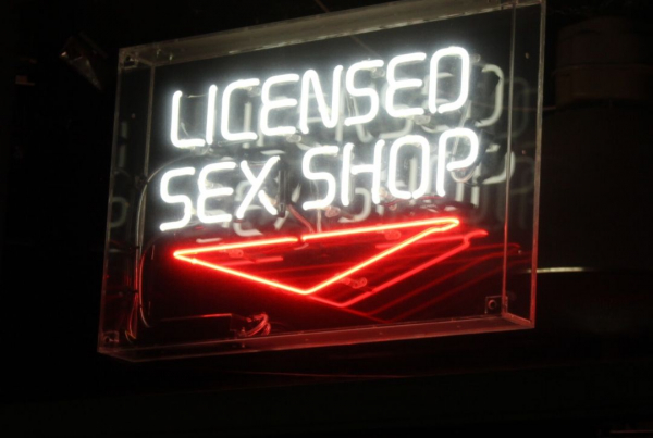 A neon sign reading 'Licenced sex shop' is lit in white against a black background, there is a red neon sign pointing downwards