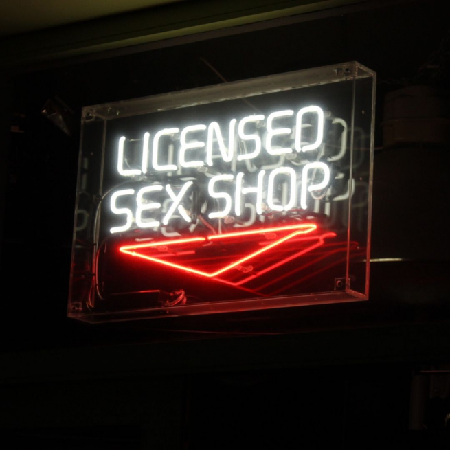 A neon sign reading 'Licenced sex shop' is lit in white against a black background, there is a red neon sign pointing downwards