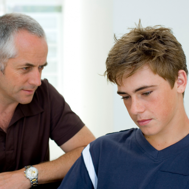 A young man is sitting to the front of the image in a blue tshirt llooking unhappy while an older man is sitting close behind him