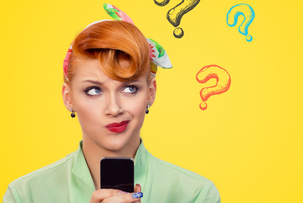 A girl stands in front of a yellow background. She has orange hair styled in a retro fashion with a headscarf and a pale green shirt. She is holding her phone in front of her and is looking off to her left with a thoughtful expression on her face. there are cartoon question marks to the side.