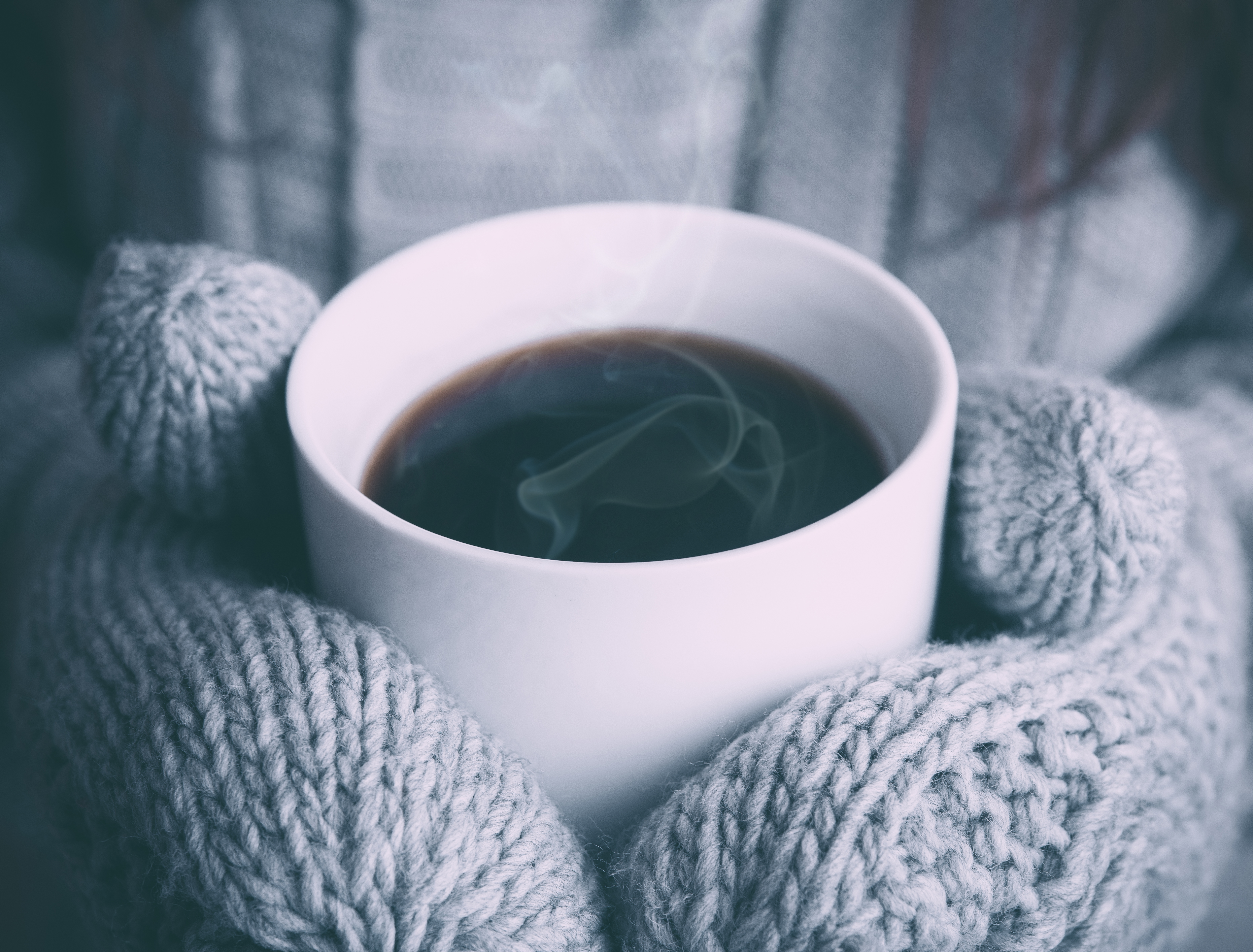 A steaming hot mug of coffee held by someone wearing knitted grey mittens