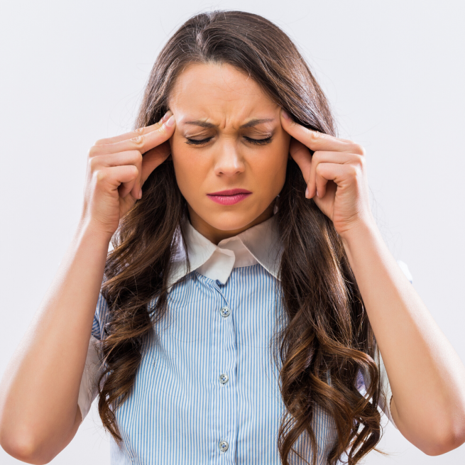 A girl with long dark hair is frowning at the camera with her hands on her temples. The is wearing a pale blue shirt with a whire collar