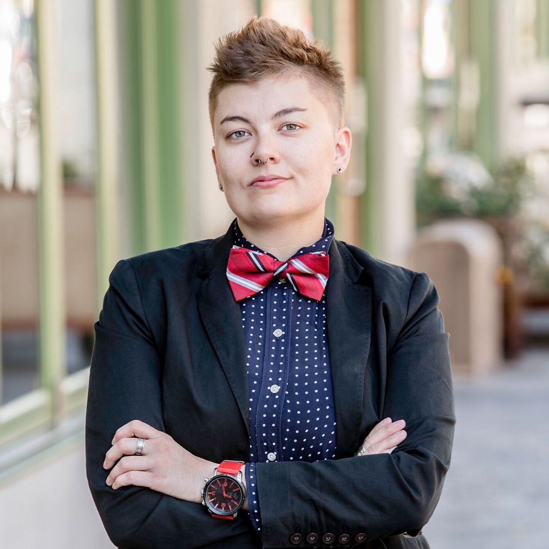 A person with short, spiked hair stands with their arms folded. They are wearing a black blazer, a navy and white polka dot shirt and a red bow tie.