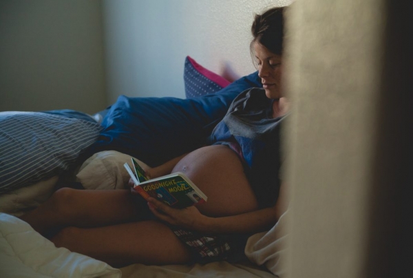 A heavily pregnant woman sitting on a bed reading a book