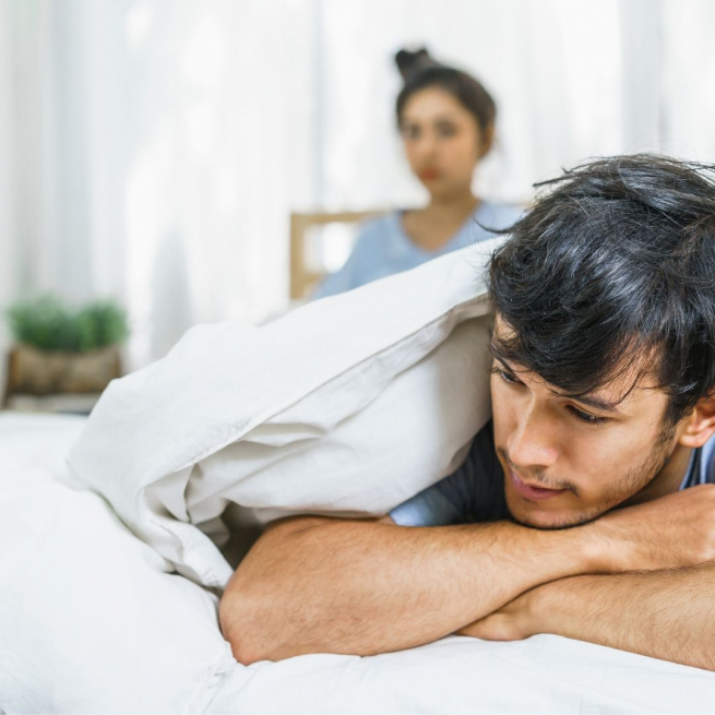 a dark-haired man is lying on his front under a white bedcover. Behind him, a woman with dark hair is out of focus