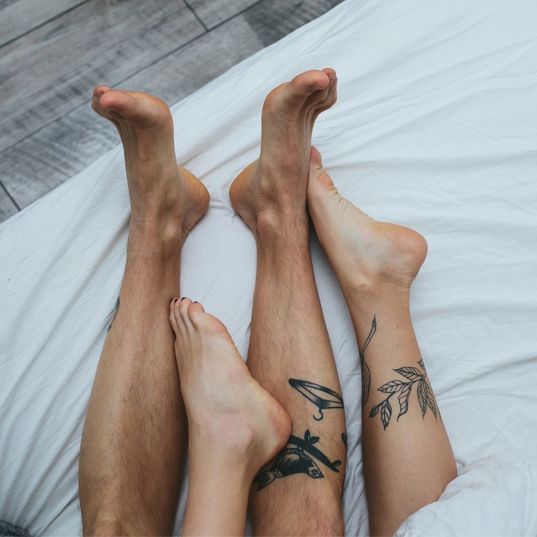 Two pairs of tattooed legs are sticking out from the bottom of a bed with white sheets