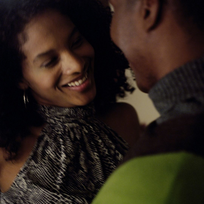 A man and woman are standing close and smiling. The woman has black curly hair and a grey halter top, the man has a jumper with a grey neckand green shoulders.