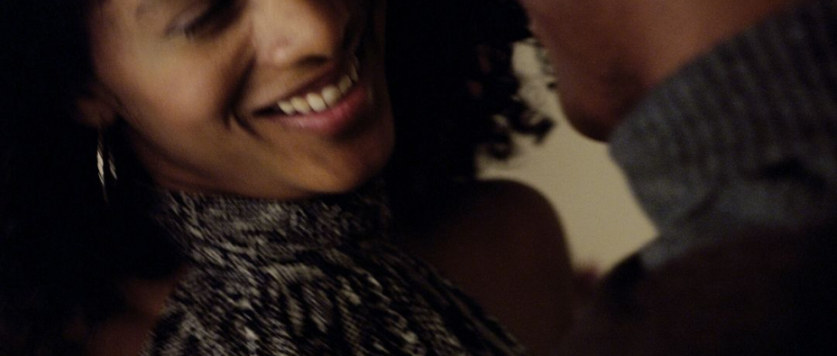 A man and woman are standing close and smiling. The woman has black curly hair and a grey halter top, the man has a jumper with a grey neckand green shoulders.