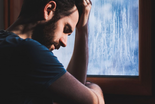 A man with dark hair and a beard looks down with a sad expression whilst leaning against a window with rain on the pane