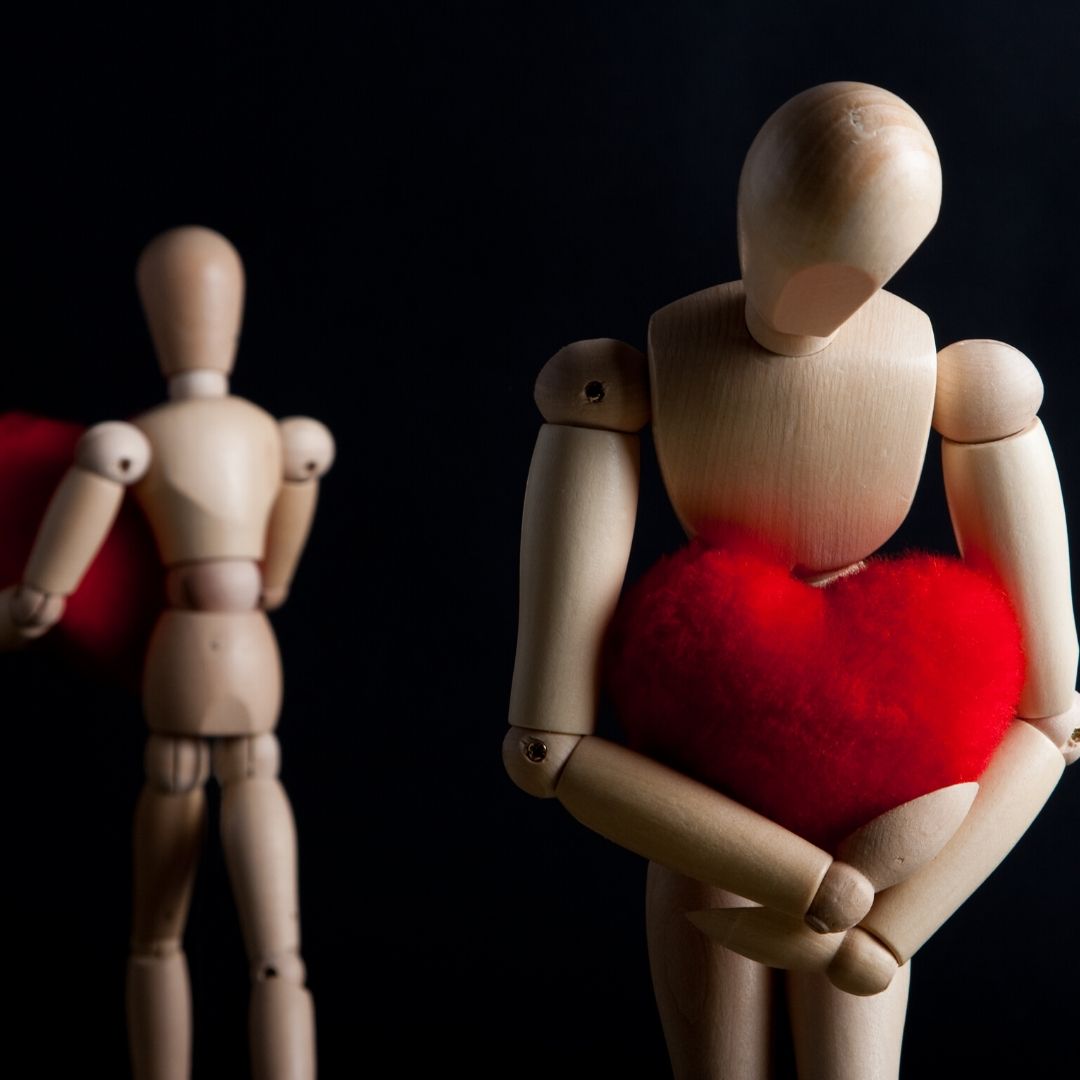 Two wooden dummies moving away from each other carrying a bright red heart