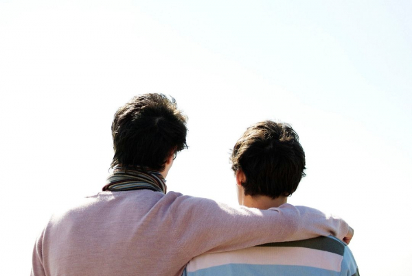 Two men looking out at the ocean with their backs to the camera, the taller one has his arm around his partner