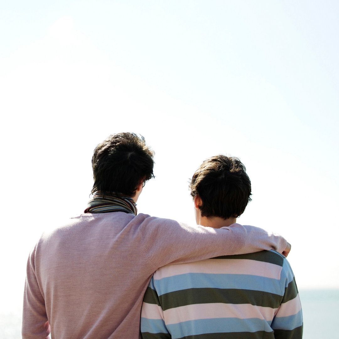 Two men looking out at the ocean with their backs to the camera, the taller one has his arm around his partner