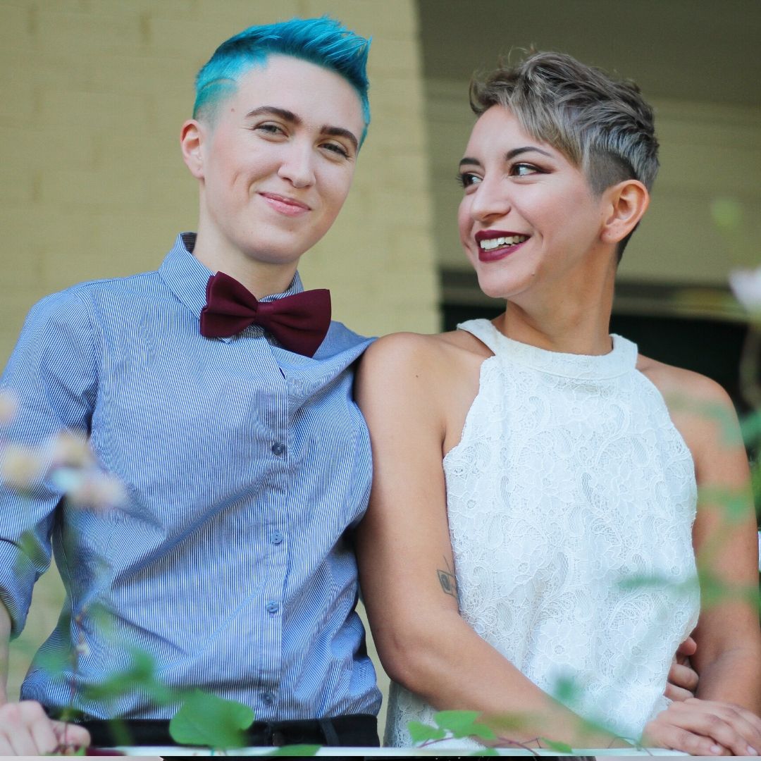 A female couple, both with very short hair. One wearing a white top, the other a blue shirt and dark red bow tie.