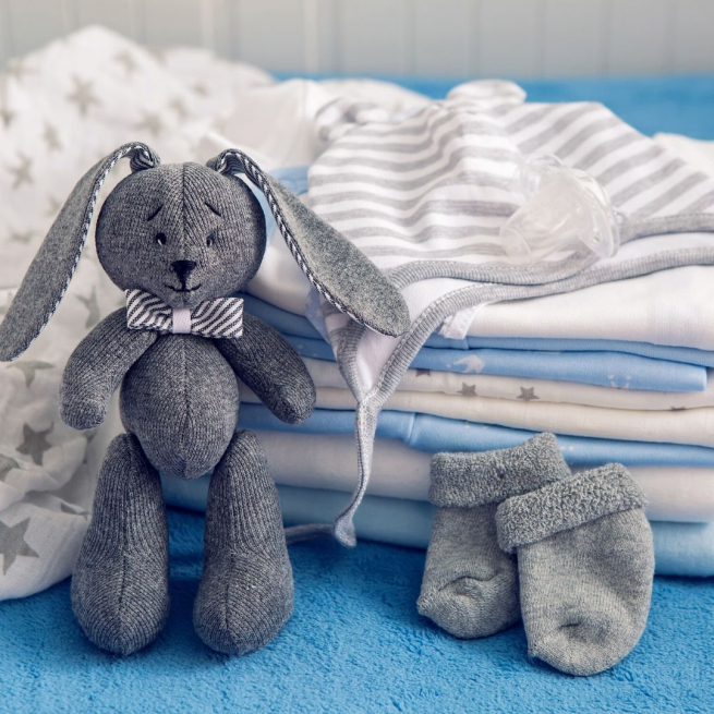A grey toy rabbit resting against a pile of baby clothes