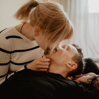 A woman leans over her partner who is lying on a bed and kisses them on the mouth, her hand rests on his chin.