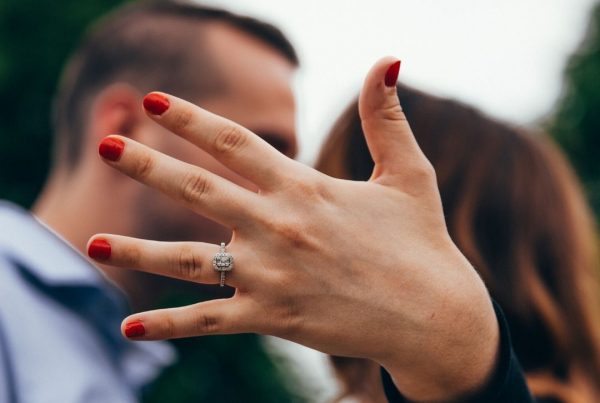 A woman holds her hand towards the camera showing her engagement ring. Blurred in the background she is kissing her fiance.