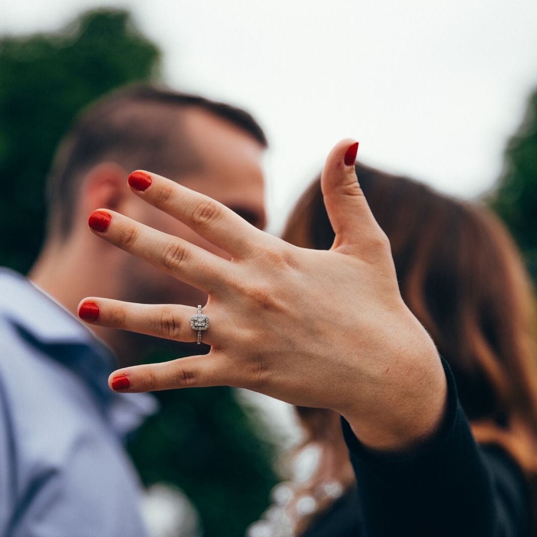 A woman holds her hand towards the camera showing her engagement ring. Blurred in the background she is kissing her fiance.