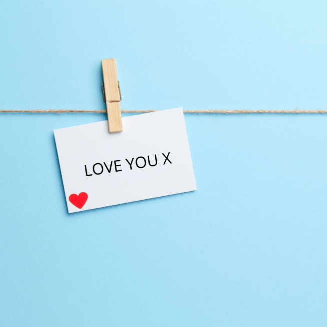 A white card with a peg holding it on a washing line. It reads 'Love You' with a red heart in the corner. The background is light blue