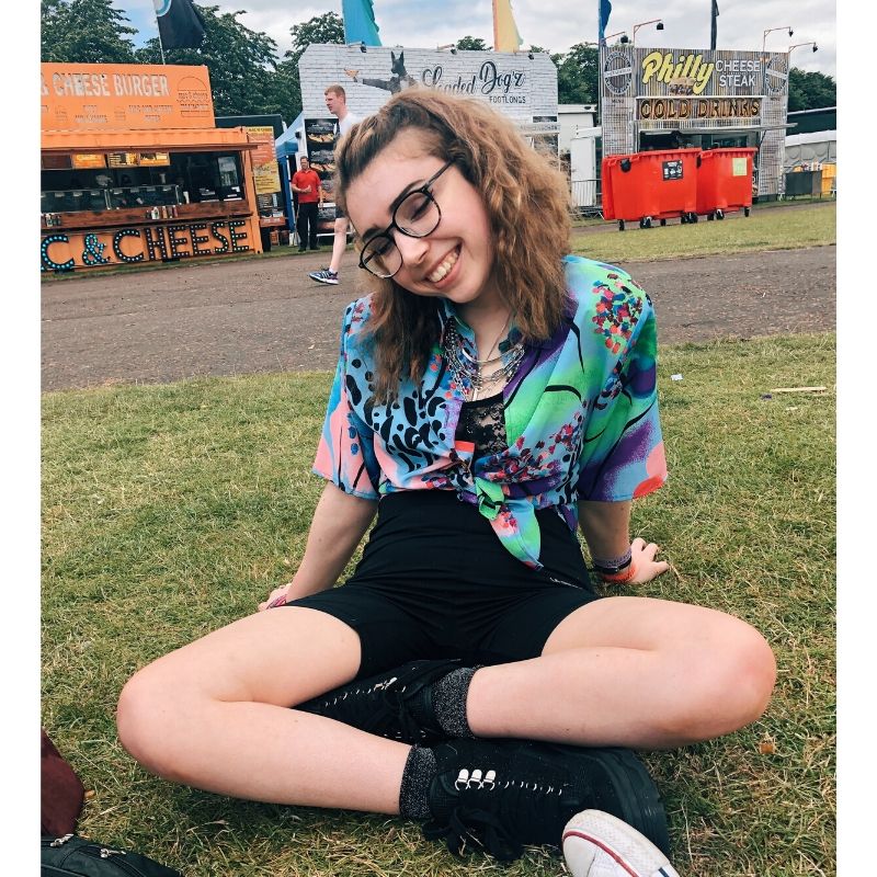 Georgia is sitting on the grass cross legged at a festival. She is wearing a pink,green and vibrant blue shirt with black shorts and boots. She is smiling and her brown shoulder length hair is wavy.