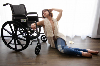 Josie on the floor leaning on her wheelchair wearing blue jeans and an open furry waistcoat