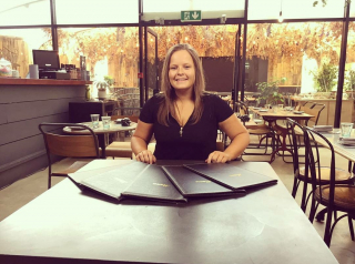 Carley Owen sits at a table in a restaurant wearing a black top and a huge beaming smile