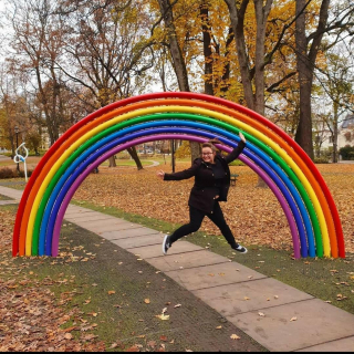 Carley is dressed in black and jumps in the air in front of a rainbow coloured archway which is situated in a park