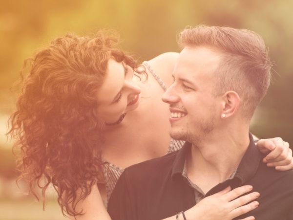 A woman with a disabled partner - she has brown curly hair leans down and puts her arms around a blonde man in a black top, they look lovingly at each other and smile