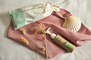 The natural love company simply lube, decoratively placed next to strands of wheat and a shell on a pink piece of fabric