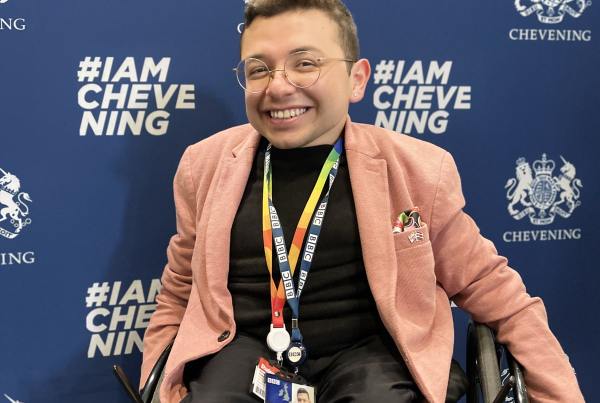 Photograph of Alex smiling at the camera in an all black outfit (t-shirt and trousers) with a pink blazor