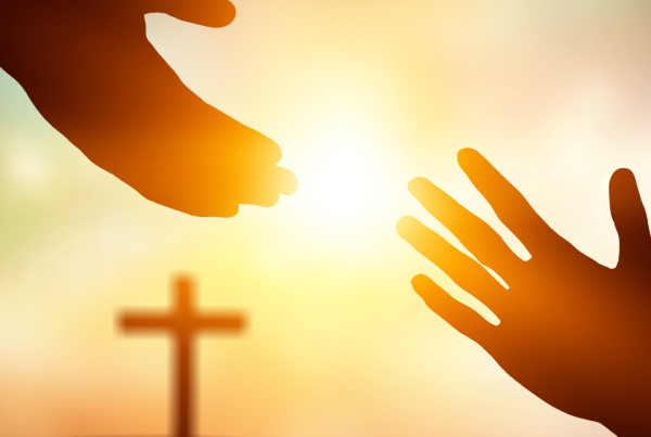 background is of a cross standing on a hill in the background with the sunsetting. In front is two hands reaching out to hold one another.