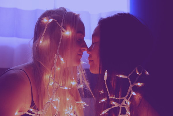 Two women sitting in bed kiss while being lit up by fairly lights to highlight how ADHD may affect your sex life.