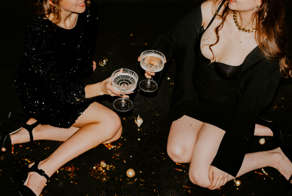 Sex positive party: two women are sitting on the floor at a party sharing a glass of champagne