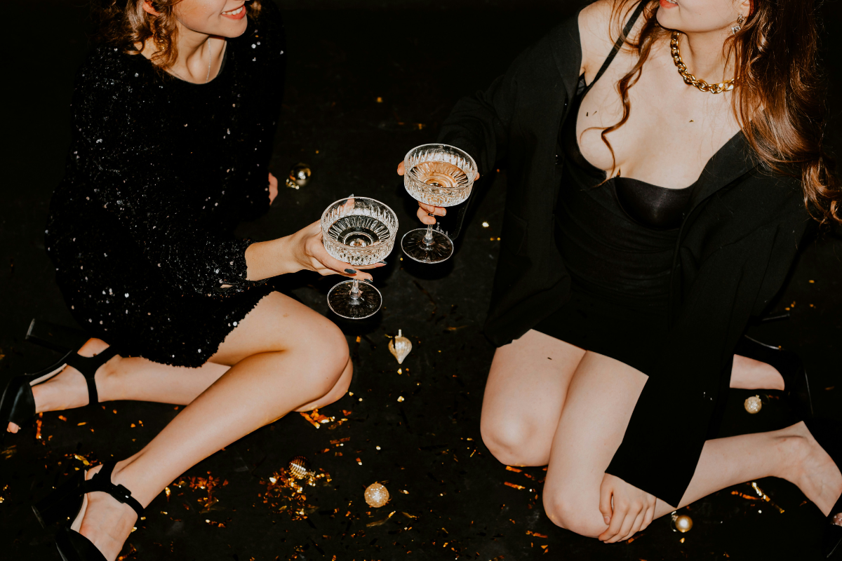 Sex positive party: two women are sitting on the floor at a party sharing a glass of champagne