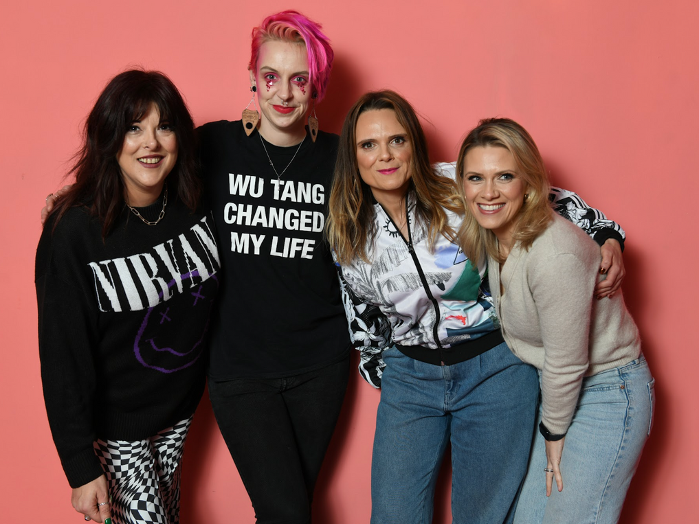 Four women posing together against a pink background, wearing band shirts and casual clothing, with one having bright pink hair.