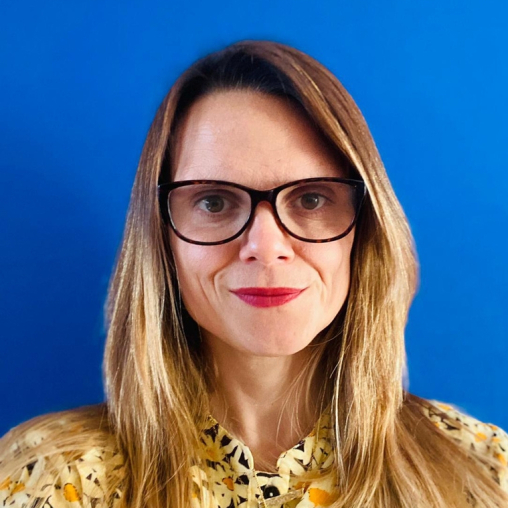 Jennie is smiling, she has long blonde hair and is wearing glasses and a yellow and black patterned top, smiling against a bright blue background.