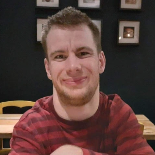 Ted is smiling. He has short brown hair and is wearing a red striped shirt, there is a table with framed pictures on the wall behind him.