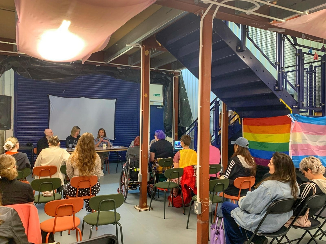 A community meeting, with diverse attendees facing speakers. A rainbow flag hangs nearby, indicating an LGBTQ+ event.