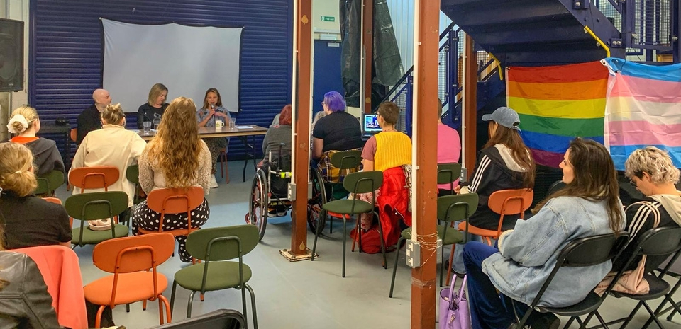 A community meeting, with diverse attendees facing speakers. A rainbow flag hangs nearby, indicating an LGBTQ+ event.