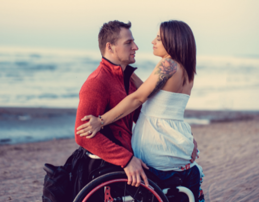 A woman sits on a man's lap. The man is sitting in a wheelchair. They are looking into each other's eyes.