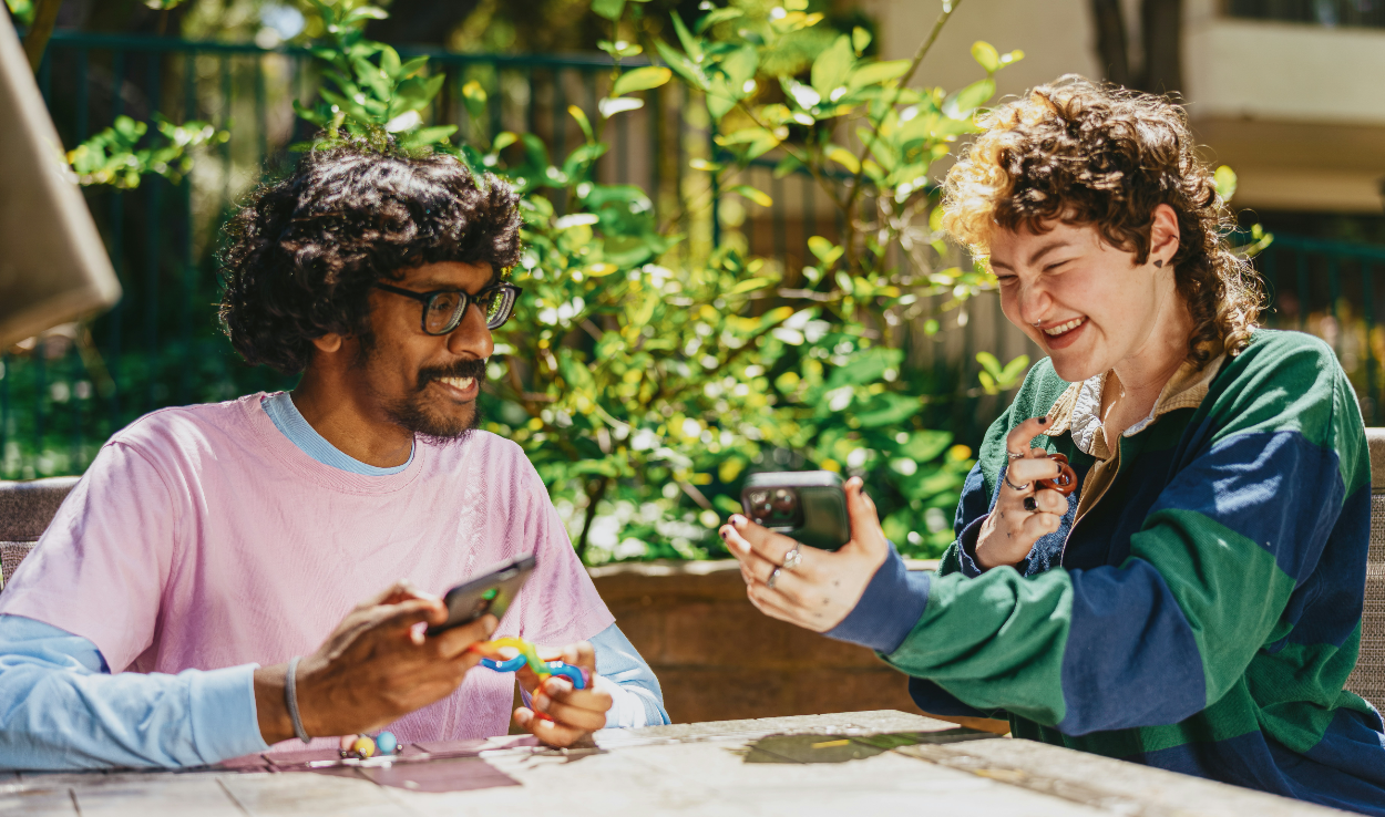 Two people conversing over a table. They are holding phones and seem to be sharing something.