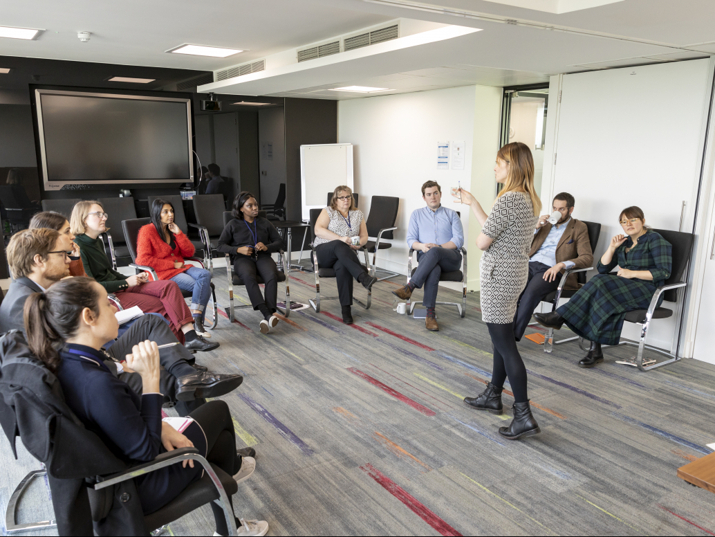 A disability awareness training session in a modern office meeting room. A woman in a patterned dress stands at the front, leading the session. About ten diverse participants sit in chairs arranged in a semicircle, listening attentively. The room features a large wall-mounted screen, a whiteboard, and colorful striped carpeting.