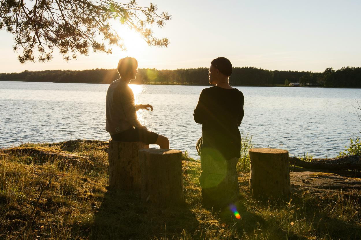 Two people conversing in front of a body of water. They are sitting on uptured logs. The sun is behind them.