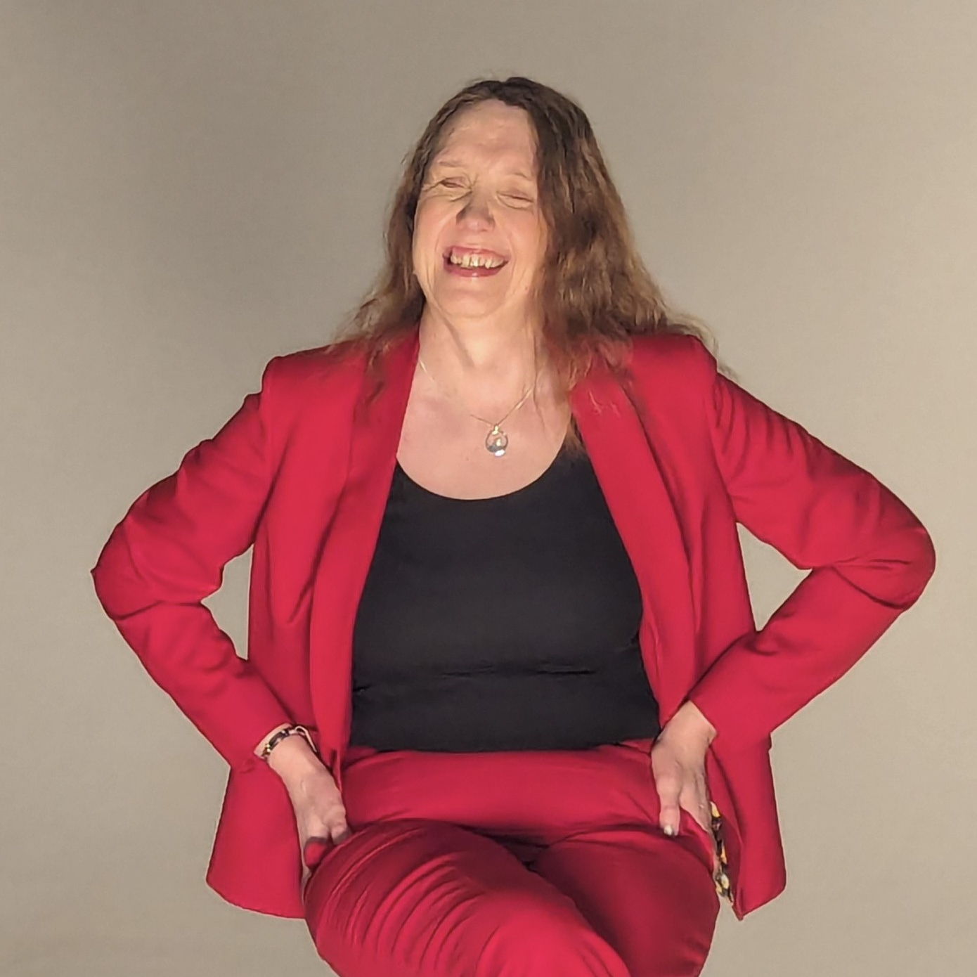 A photograph of Sarah Stephenson-Hunter. She is sitting on a stool. She is wearing a red jacket and trousers, with a black top. She is smiling.