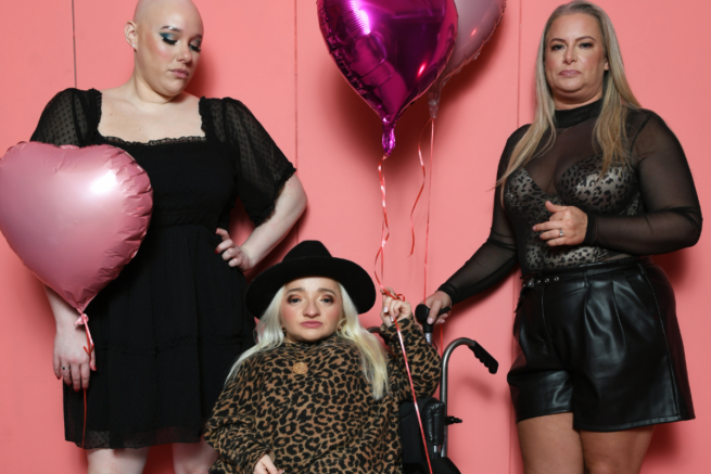 Three people are in a group: one person is wearing a black dress and holding a pink heart balloon, another is sitting in a wheelchair wearing a leopard print top and black hat. The other person is standing wearing all black