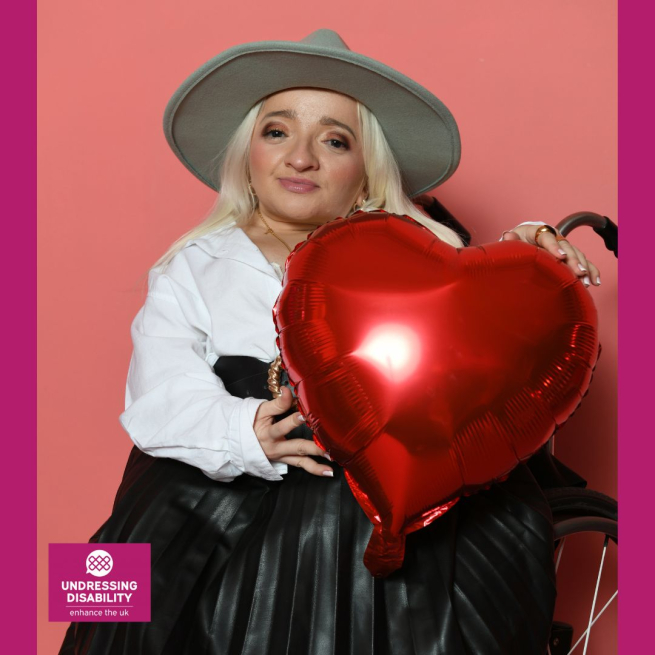 Sam has long blonde hair and is wearing a light green hate. She is sitting in a wheelchair holding a red shiny balloon shaped like a heart. She wears a white shirt and black skirt