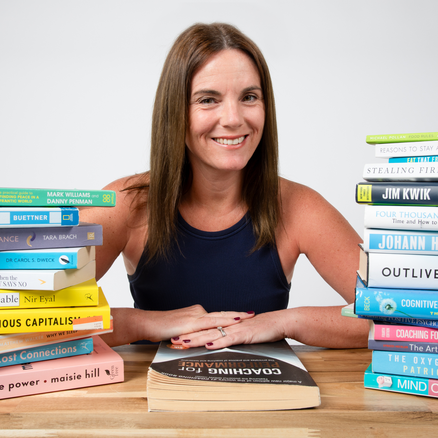 Michelle Flynn sitting at a desk. She has a stack of books on either side, and is laying her hands on a book on the table titled "Coaching for Performance". She is smiling.