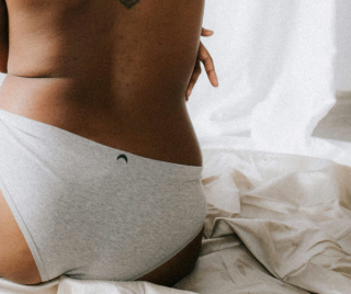 Periods: a person sitting with their back to the camera. They are not wearing a top but they are wearing grey underwear. They are sitting on a bed