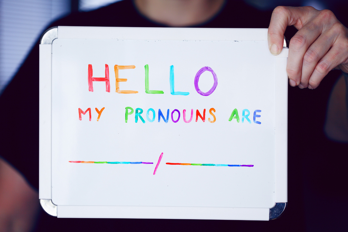 Identity: a person holding a white sign that has 'hello my pronouns are......' written in rainbow pen.